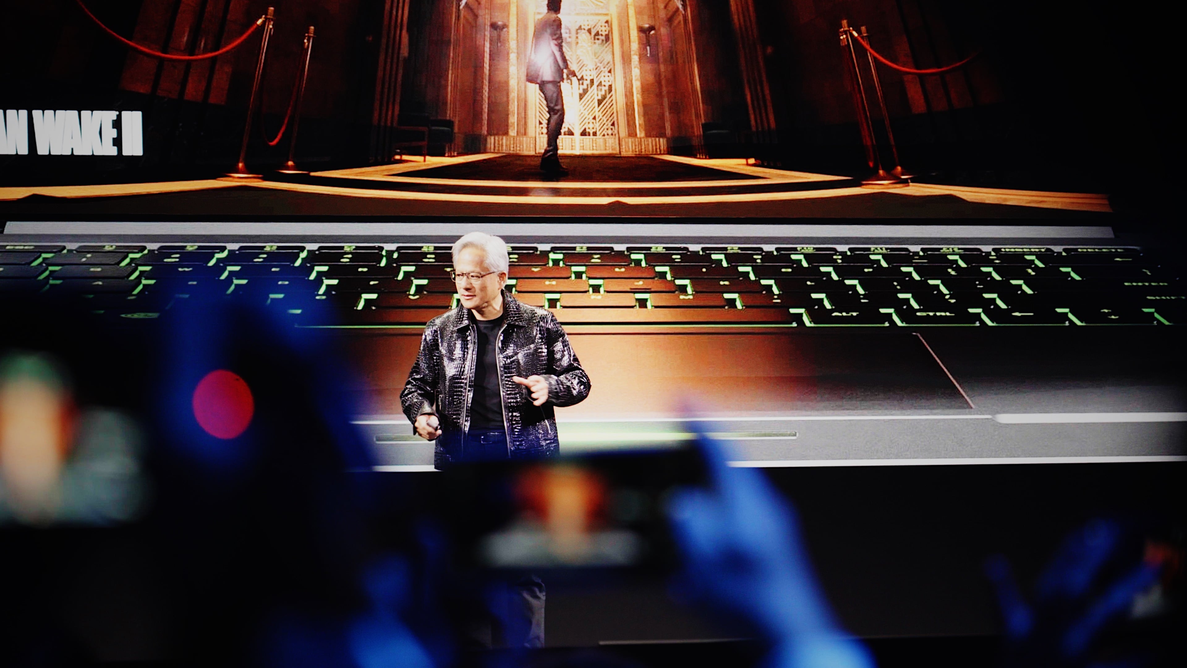 A man in a leather jacket and glasses speaks onstage in a large arena.